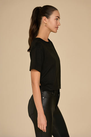 A person with long brown hair in a ponytail is wearing a Naya Suede T-Shirt in black with drop sleeves and black pants, standing in profile against a beige background.