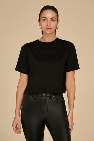 A person with tied-back hair is wearing a black Naya Suede T-Shirt in a cropped fit, paired with black leather pants, and standing against a beige background. They have a neutral expression and are accessorized with hoop earrings, highlighting the versatility of the piece for any occasion.