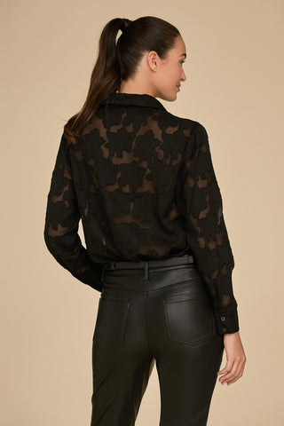 A woman with a ponytail is facing away, showcasing the Stelle Lace Blouse in black, paired with black leather pants. The blouse features lightweight lace and floral patterns. The soft beige background creates a striking contrast with her dark ensemble.