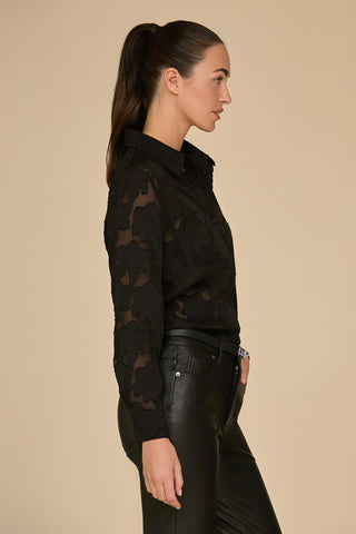 A woman with long dark hair in a ponytail is wearing a black Stelle Lace Blouse and black leather pants. She stands in profile against a plain beige background, the lightweight lace adding an elegant touch to her sleek, stylish outfit.