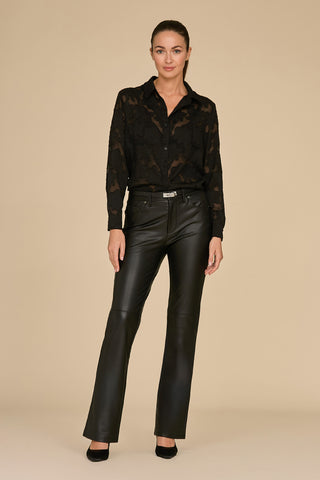 A person wearing a sheer black blouse similar to the Stelle Lace Blouse - Black, along with sleek black leather pants, stands against a beige background. Their hair is pulled back, and they're stylishly perched in black heels.