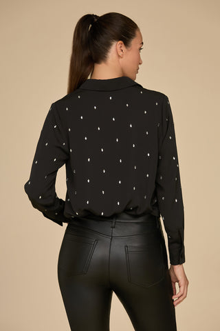 A woman with long brown hair in a ponytail is facing away, wearing the Aniya Crystal Shirt by Noli Yoga—a statement-style black blouse featuring a small white dotted pattern—and black leather pants. The background is plain beige.