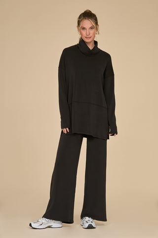 A person stands against a beige background in a Daphne Pant - Black and wide-leg pants, paired with white sneakers. Crafted from a soft modal blend, the ensemble offers luxurious comfort as they look directly at the camera with a relaxed expression.