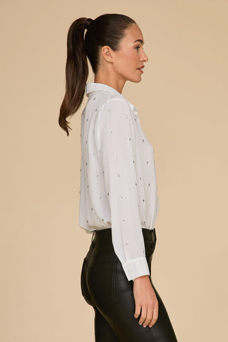 A woman with a ponytail is wearing the Aniya Crystal Shirt in white, featuring a delicate diamond pattern, paired with black leather pants. She stands sideways against a plain beige background, exuding effortless elegance.