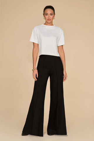 Against a beige background, a person stands wearing an ivory Naya Suede T-Shirt with a cropped fit and black wide-leg pants. Their hair is tied back, complemented by gold hoop earrings and a bracelet. The overall look is minimalist and chic, perfect as a stylish layering piece.