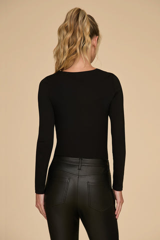 A person with wavy blonde hair in a ponytail is facing away from the camera. They are wearing the curve-flattering Jada Cut Out Long Sleeve Top in black and black leather pants, set against a plain beige background.