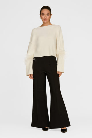 A person poses against a plain backdrop wearing the Bella Feather Trim Sweater in Pearl, featuring feather-detailed sleeves, paired with black flared pants, black shoes, and gold earrings. Their hair is styled back neatly.