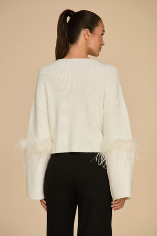 A woman with a ponytail is wearing the Bella Feather Trim Sweater in pearl, featuring feather accents on the sleeves, paired with black pants. She is standing with her back to the camera against a beige background.