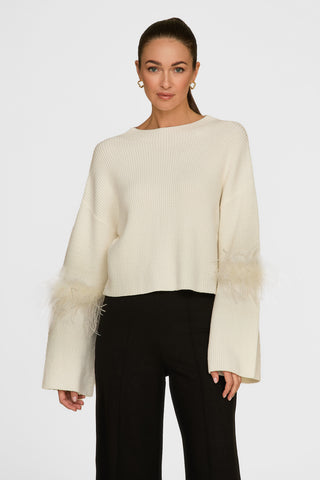 A woman dons the elegant Bella Feather Trim Sweater in Pearl, complemented by black pants. The sweater's feathery sleeve details enhance its sophistication against a simple white background.