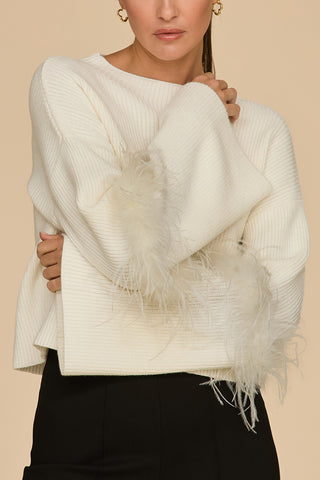 A person wearing the Bella Feather Trim Sweater - Pearl, featuring luxurious feather details on the sleeves, paired with sleek black pants. The contemporary ensemble stands out brilliantly against the light beige background.