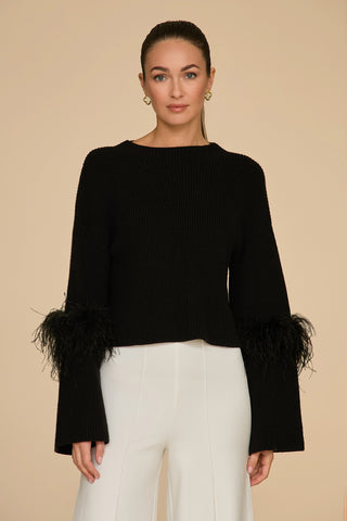 A woman poses against a beige backdrop, dressed in the Bella Feather Trim Sweater in black, paired with white pants. She accessorizes with gold hoop earrings and her long hair is tied back.