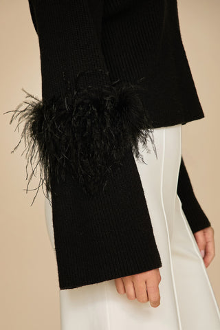 Close-up of a person wearing the Bella Feather Trim Sweater in black, paired with white pants. The textured feather detail adds a stylish and dramatic flair to the outfit against a plain beige background.