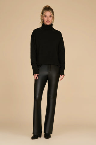 A person stands against a beige background, wearing the oversized Gia Turtleneck Sweater in black and black leather pants. Their hair is styled in a loose updo, and they are wearing black shoes.