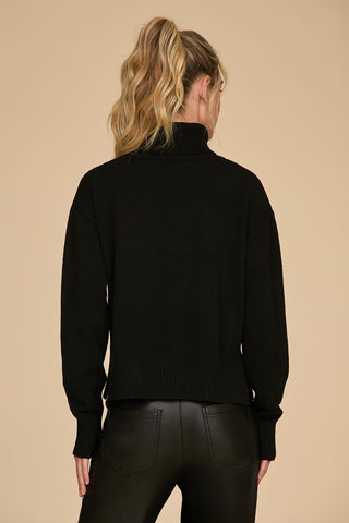 A person with wavy blonde hair in a ponytail is wearing the oversized Gia Turtleneck Sweater in black along with black leather pants, facing away. The background is a solid beige color.