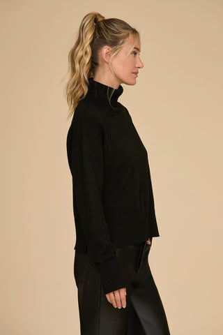 A person with long, wavy hair tied in a ponytail is wearing the oversized Gia Turtleneck Sweater in black paired with matching pants, standing in profile against a beige background and exuding a chic and cozy vibe.