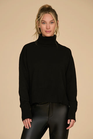 A person with long hair is wearing the Gia Turtleneck Sweater in black alongside a pair of black leather pants, posing against a neutral beige background. With a relaxed smile and arms slightly bent, they exude effortless style in this chic ensemble.