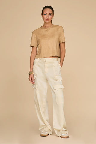 A person stands against a beige background, wearing a short-sleeved tan top and the Milan Satin Cargo Pant in pearl, showcasing exquisite cargo detail. With one hand in their pocket and bracelets adorning their wrist, they add effortless style to this versatile look.