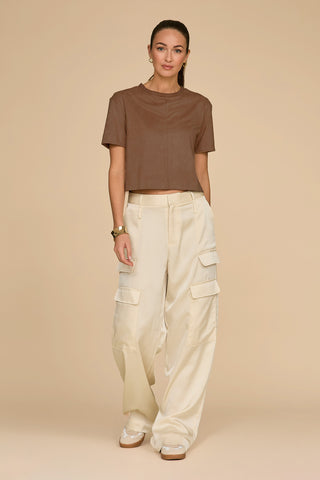 A person stands on a beige background, wearing the Naya Suede T-Shirt in Java with a cropped fit and cream cargo pants. They have long hair tied back, wear hoop earrings and a bracelet, and have their hands casually by their sides.