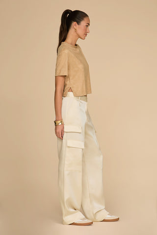 A person stands in profile against a beige background, wearing a tan short-sleeve top and the Milan Satin Cargo Pant in Pearl, which features versatile cargo details. They have brown hair tied back and accessorize with gold earrings, a bracelet, and white sneakers.