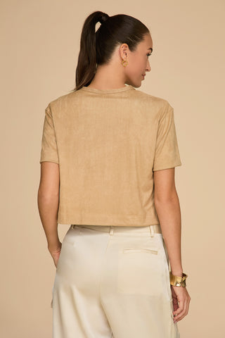 A woman with a ponytail stands facing away, dressed in a Naya Suede T-Shirt in Cafe au Lait and light-colored trousers. Her gold hoop earrings and wristwatch enhance the elegance against the simple, neutral background.