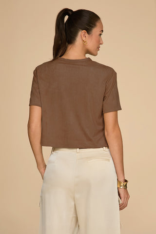 A person with long hair styled in a ponytail is wearing the Naya Suede T-Shirt in a cropped fit, paired with light-colored pants. They are standing against a beige background, facing away from the camera, and accessorized with gold earrings and a bracelet.