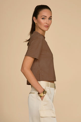 A woman with long brown hair in a ponytail stands in profile against a beige background, wearing the Naya Suede T-Shirt - Java with a cropped fit and white pants featuring pockets. Her hands rest casually in her pockets as she accessorizes with earrings and a bracelet, creating the perfect layering piece.