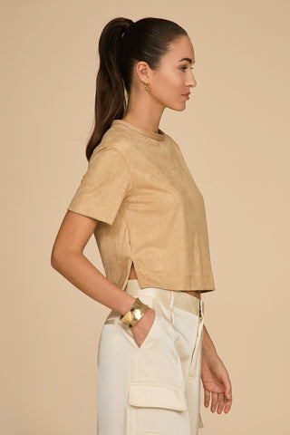 A woman with a high ponytail stands in profile against a beige background. She is wearing the Naya Suede T-Shirt in Cafe au Lait paired with cream-colored cropped pants, one hand casually placed in her pocket. Her ensemble is complemented by gold hoop earrings and a gold bracelet.