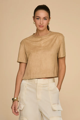 A person with long hair tied back, wearing a Naya Suede T-Shirt in Cafe au Lait and light cargo pants, stands against a beige background. Their cropped fit is perfectly accented with hoop earrings and a bracelet.