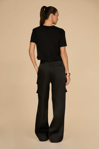 A woman with long hair tied in a ponytail stands facing away, showcasing her sleek black t-shirt paired with the Milan Satin Cargo Pant in black. The neutral beige background subtly highlights the chic ensemble's versatility.