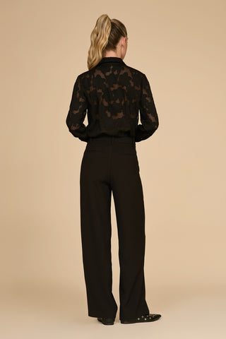 A confident person with blonde hair in a ponytail stands stylishly with their back to the camera against a beige background, wearing a sheer black blouse and the Lia Pleated Trouser in black. Their fashionable black shoes are adorned with small decorative studs.