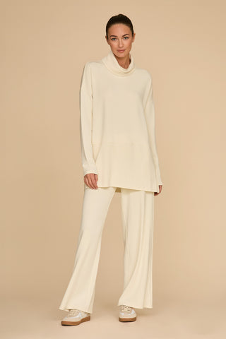 Against a beige background, an individual stands wearing a cream-colored ensemble called the Daphne Pant - Pearl, which includes a long-sleeve oversized top and wide-leg pants crafted from a silky, breathable soft modal blend. Their brown hair is pulled back, and they're sporting white sneakers with brown soles for the perfect on-the-go look.