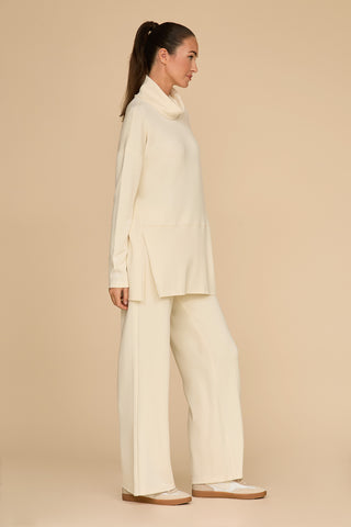 A person with long hair styled in a ponytail showcases the ultimate on-the-go look in a cream-colored outfit. The loose turtleneck top paired with the Daphne Pant - Pearl, made from a soft modal blend, exudes comfort as they stand sideways against a plain beige background.