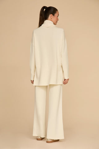 A person with long hair tied back is standing and facing away, showcasing the ultimate on-the-go look in a cream-colored turtleneck top paired with the Daphne Pant in Pearl. This outfit, crafted from a silky and breathable soft modal blend, beautifully complements the light beige background.