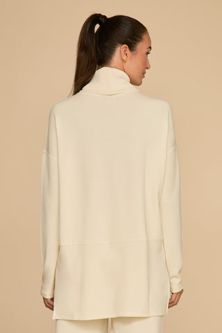 The image depicts a person with long hair tied in a ponytail, wearing the Daphne Top in Pearl, made from a soft modal blend cream-colored turtleneck and matching pants, facing away from the camera against a plain background—perfecting the ultimate on-the-go look.