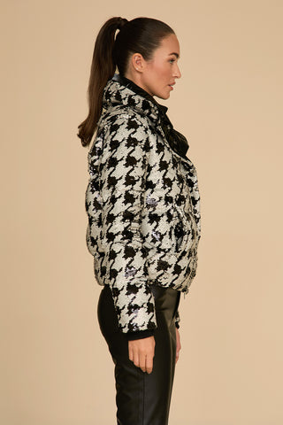 A woman with a ponytail stands in profile against a beige background, exuding elegance in her high-collared Sequin Puffer Jacket - Houndstooth paired with black leather pants—a stylish blend perfect for any winter wardrobe.