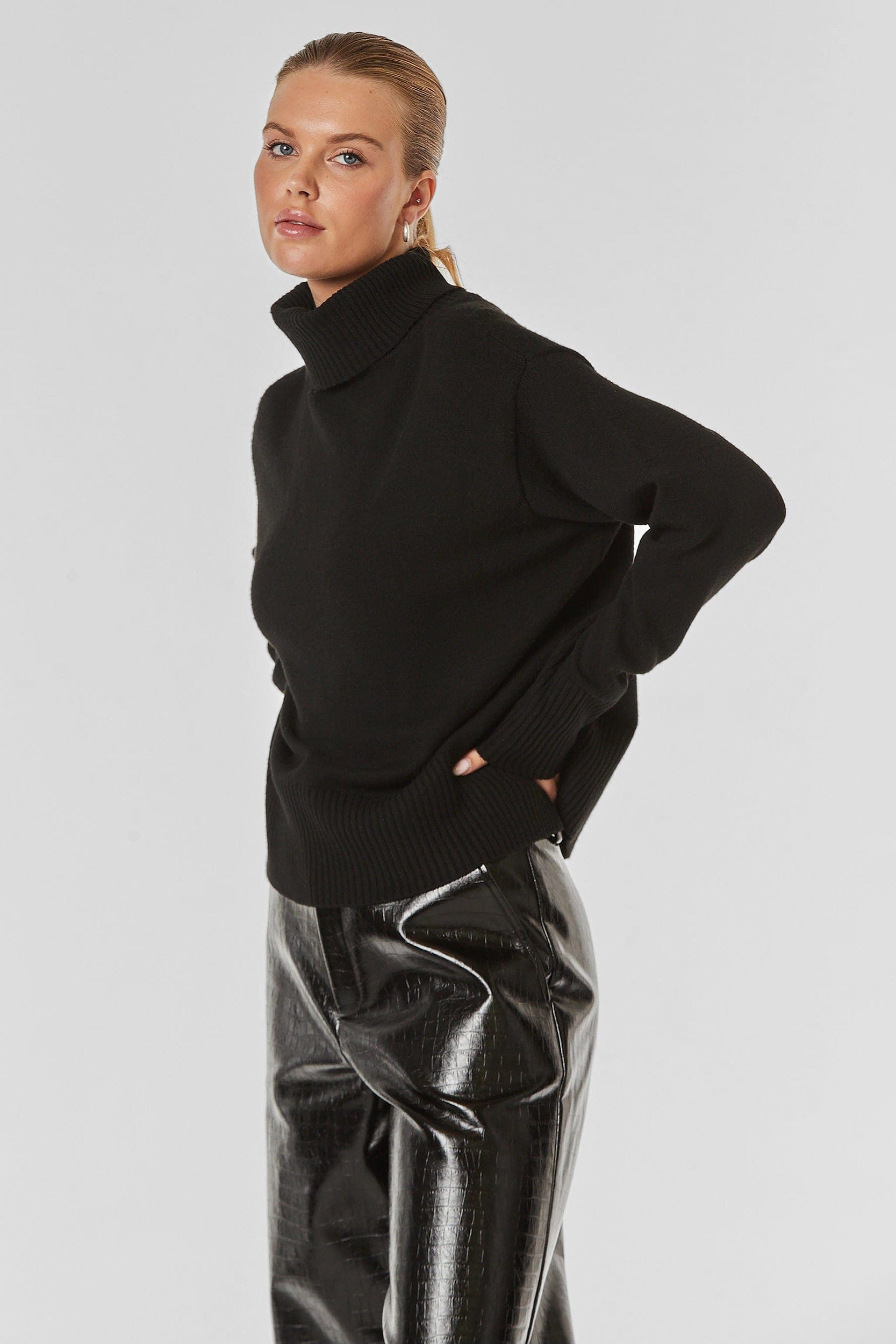 A person with blond hair is wearing a black Gia Oversized Sweater and black vinyl pants. They have their hands in their pockets and are looking directly at the camera against a plain grey background.