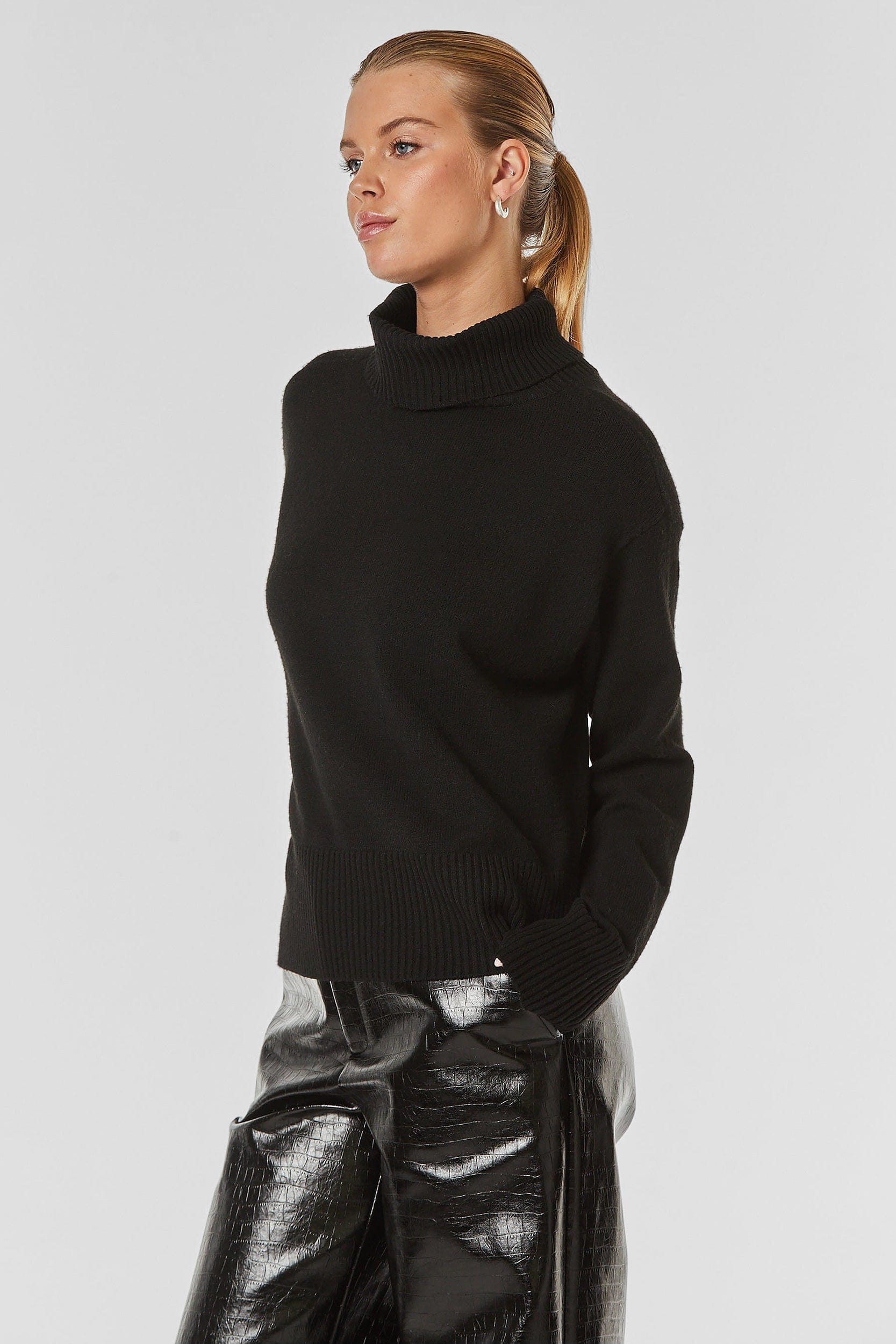 A person with blonde hair tied back in a ponytail is wearing the Gia Oversized Sweater in black and shiny black pants. They are standing against a plain grey background with one hand in their pocket, looking to the side.