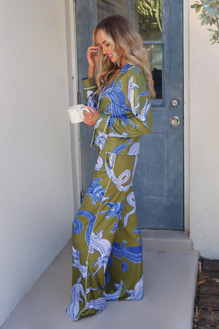 A woman with long, wavy hair stands on a porch in front of a blue door, wearing a matching olive green pajama set with Nile gator print and the Nile Pajama Soft Long Pant. She holds a white speckled coffee mug in one hand and touches her hair with the other, smiling.