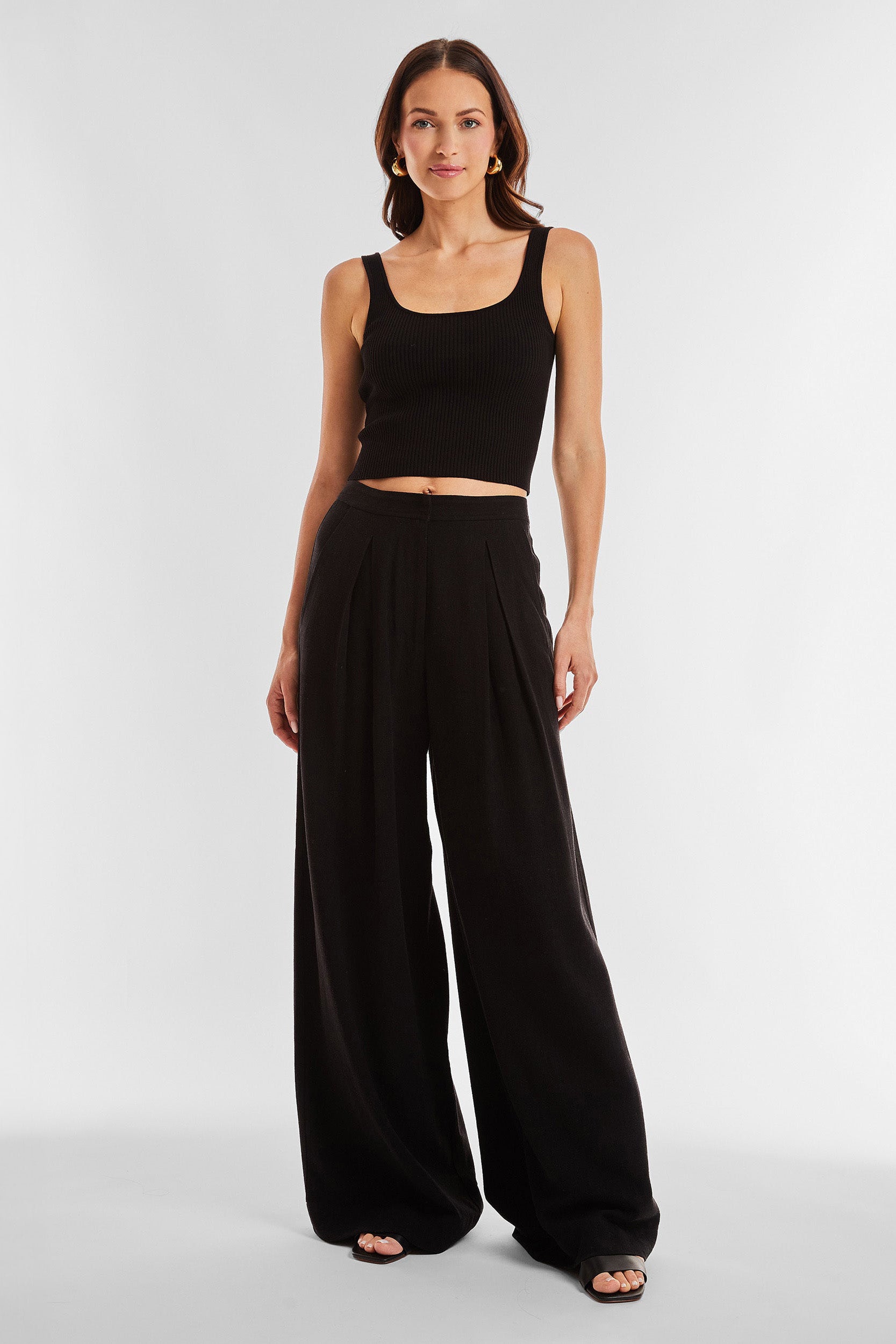 A woman with long hair wearing a black sleeveless crop top and the Seychelles Relaxed Linen Pant in black, designed with a high-waisted, wide-leg fit, exudes confidence against a plain, light-colored background. She completes her look with open-toed black shoes.