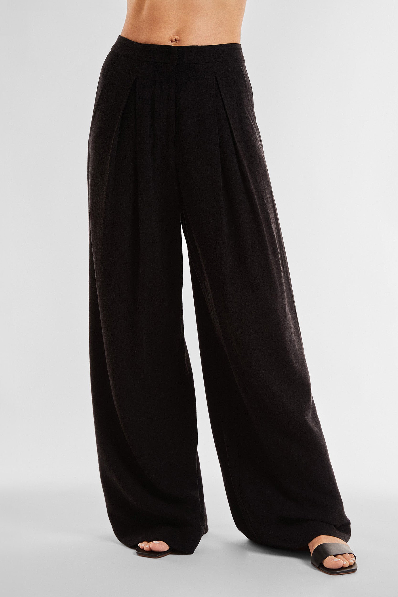 A person is modeling the Seychelles Relaxed Linen Pant - Black, which features wide legs and front pleats. These high-waisted, relaxed linen pants offer a breathable fit and are paired with black open-toe sandals. The photo is taken against a plain white background, showcasing only the model's lower torso and legs.