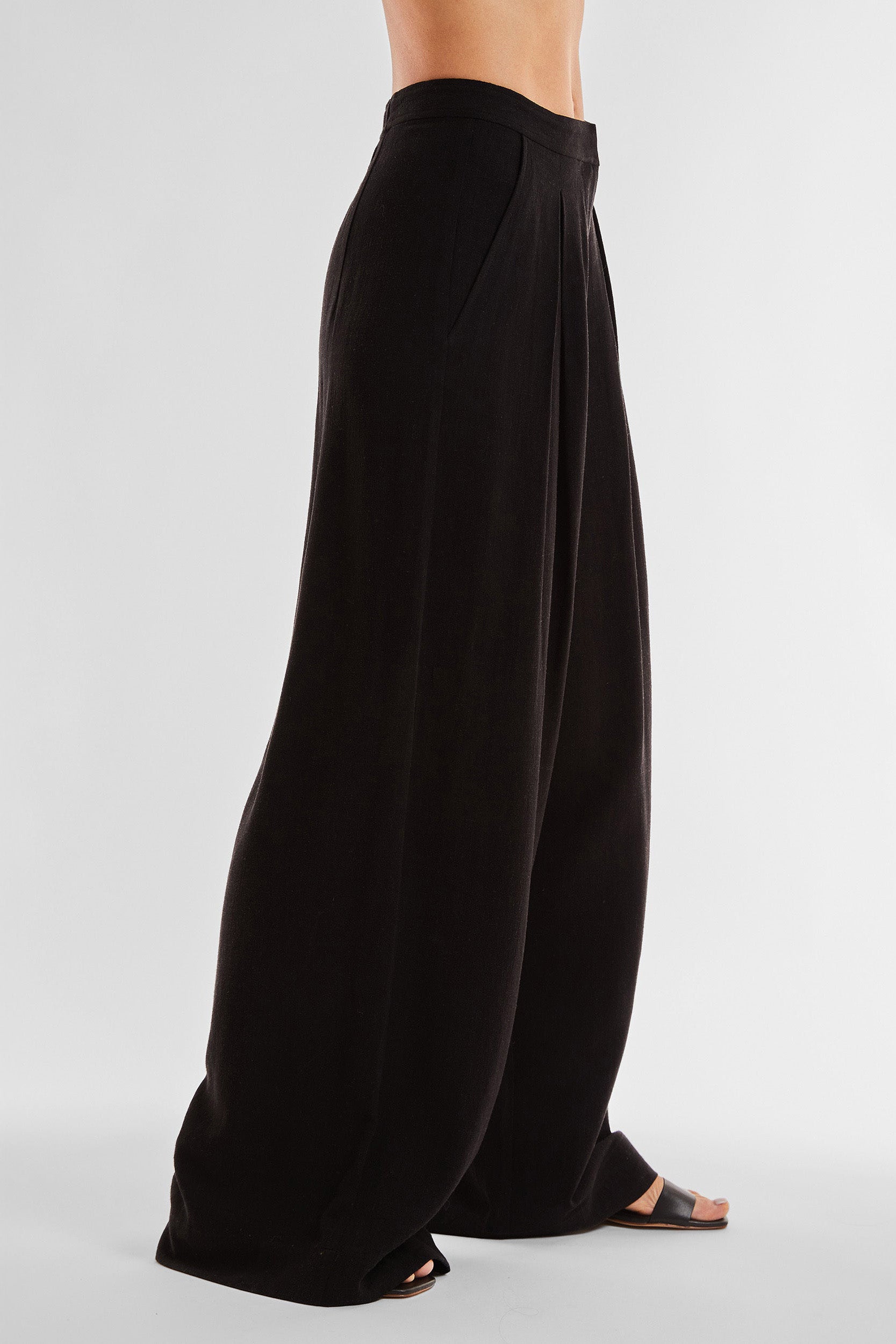 A person is wearing the Seychelles Relaxed Linen Pant - Black, which features a high-waisted, wide-leg design with pleats. The pants have a loose, flowing fit and are paired with black open-toed sandals. The image shows a side view of the lower half of the person's body against a plain background.