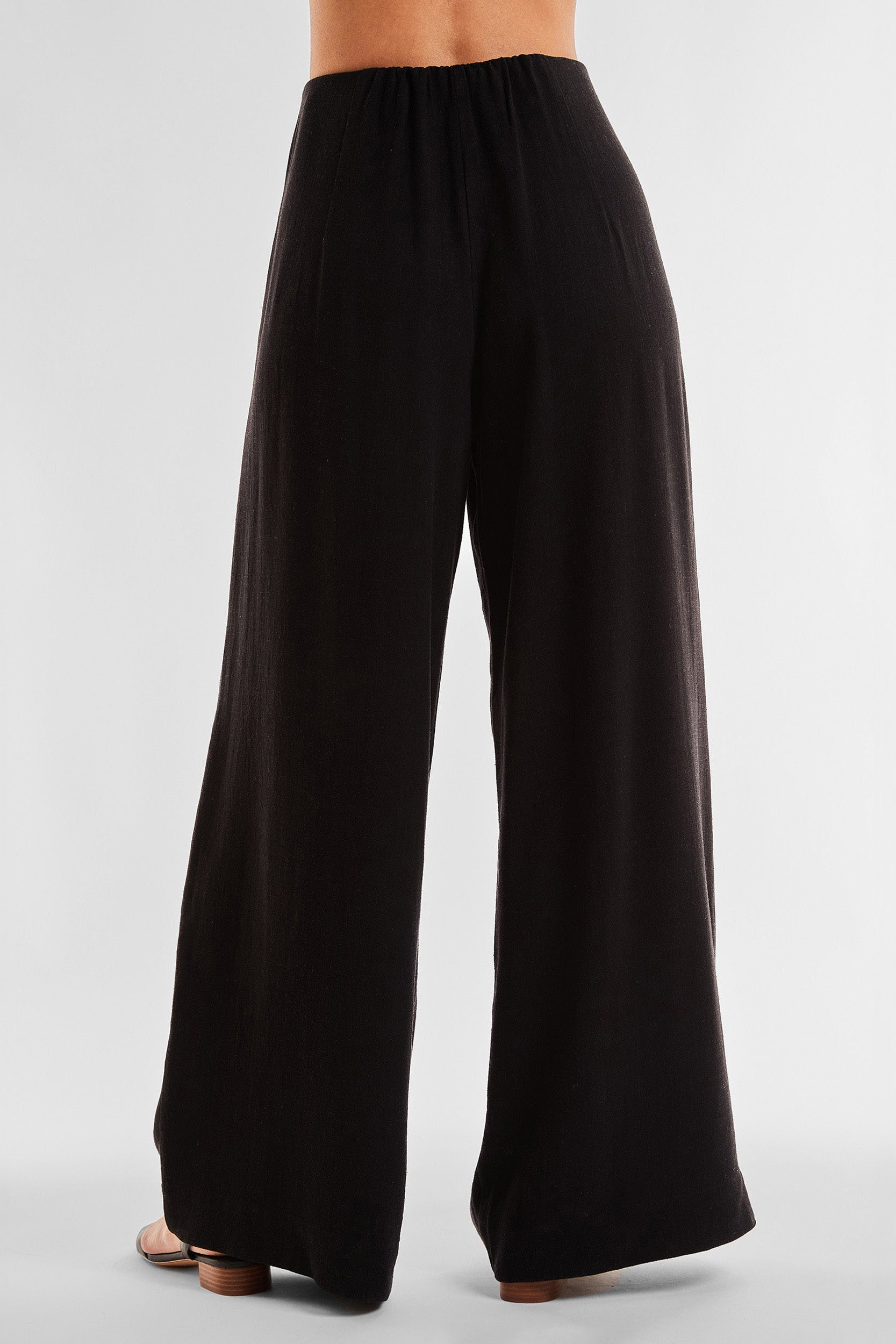 A person is shown from the waist down, wearing the Seychelles Relaxed Linen Pant - Black. The high-rise, wide-leg pants are made of a lightweight fabric and the person is standing against a plain white background.