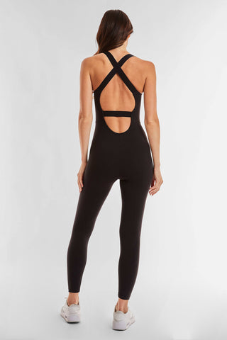 A woman in the squat-proof Vixen Jumpsuit - Black, featuring a crisscross open-back design, stands with her back to the camera. Her long brown hair falls gracefully down her back, enhancing the flattering fit of her outfit. She completes her look with white sneakers against a simple white background.