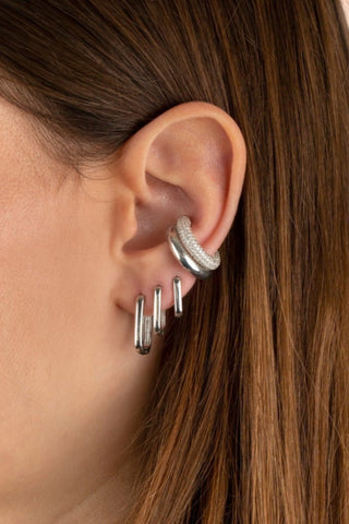 Close-up of a person's ear adorned with multiple sterling silver earrings. The striking combination includes two thick hoop earrings in the helix (upper ear) and three elegant Solid Oval Huggie Earrings by By Adina Eden in the lobe, creating a stylish and modern look. The person has long, straight brown hair.