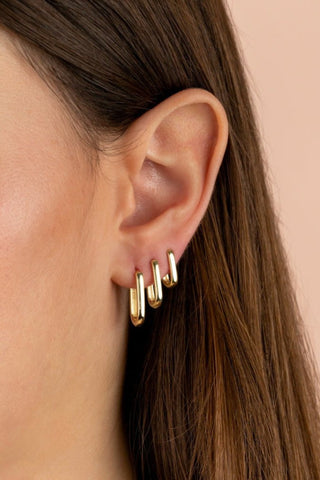 Close-up of a person's ear adorned with four small, Solid Oval Huggie Earrings by By Adina Eden along the outer edge. The background is a soft, solid peach color. The person's brown hair partially covers the ear.