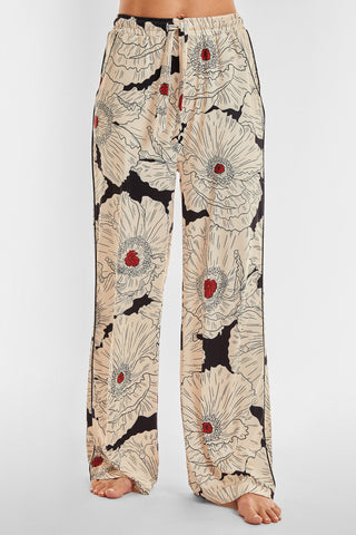 A person is wearing the Poppy Pajama Soft Long Pant, featuring a loose fit in cream with an eye-catching large black and red floral poppy print. The high-waisted pants have a drawstring waist and wide legs, revealing their bare feet against a plain white background.