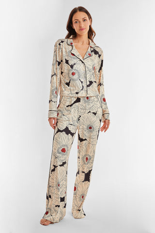 A woman stands against a plain white background, wearing the Poppy Pajama Soft Long Sleeve Shirt adorned with a large floral Poppy print featuring beige flowers and red centers, paired with loose-fitting pajama pants. She is barefoot.