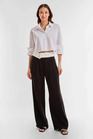 A woman with long, dark hair wears a white, cropped button-up shirt with rolled sleeves. She pairs it with high-waisted, loose-fitting Soho Foldover Trousers in black from Noli Yoga that feature a distinctive white waistband. The ensemble is completed with black sandals. She stands against a plain white background, creating an impactful style statement.