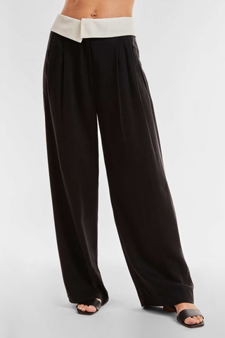 A person is modeling the Soho Foldover Trouser - Black by Noli Yoga, featuring a stylish white waistband. These black wide-leg trousers, crafted from lightweight fabric, are paired with black open-toe sandals. The plain white background accentuates the trousers' design and fit, making them a standout statement piece. The model's upper body is not visible.