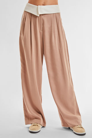 A person wearing Noli Yoga's Soho Foldover Trouser in Mocha, featuring a white waistband, stands against a plain background. The versatile spring trousers, designed with pleats and a relaxed fit, drape down to touch beige sneakers. The lightweight fabric ensures comfort, though the upper body is not visible in the frame.